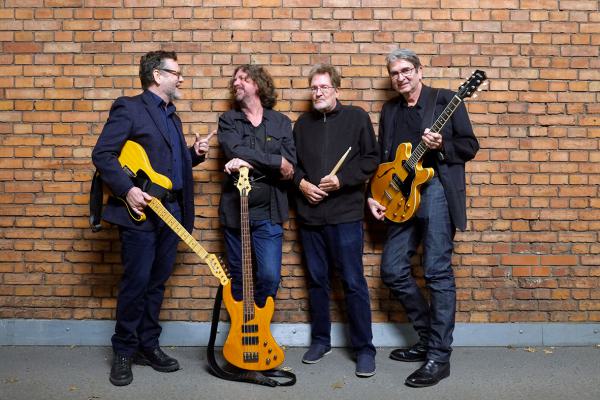 Bandfoto The Velvet Suits: Vier Männer stehen locker vor einer Backsteinwand. Zwei von ihnen halten E-Gitarren, einer hält einen Bass, und der vierte hat Drumsticks in der Hand. Alle lachen oder unterhalten sich miteinander und wirken entspannt und gut ge